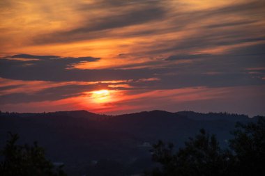 Bu görüntü, karanlık gökyüzüne karışan sıcak renklerle, dağlık bir arazide canlı bir gün batımı yakalar. Ufuk dağlarla dolu.