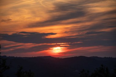 Canlı bir günbatımı ufku aydınlatır, sıcak, turuncu bir parıltı yayar, uzak tepeleri gölgeler içinde siluete boğar..