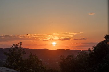 Alacakaranlıkta yakalanan güneş, yuvarlanan tepelerin ardından iniyor. Gökyüzünü canlı portakallar ve sarılarla boyuyor. Ağaçların silueti ve uzak tepeler.