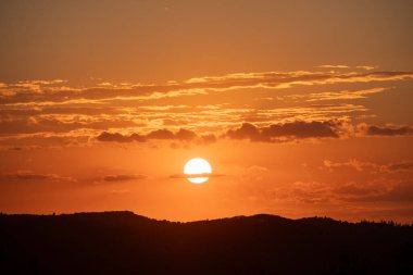 Güzel turuncu bir gün batımı siluetli bir manzaranın üzerinde, bulutların arasından parlayan altın ışıkla. Gökyüzü canlı ve renkli bir başyapıttır..