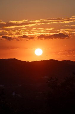 Batan güneş turuncu bir parıltı yayıyor, siluet gibi görünen dağlar ve ağaçlar. Bulutlar turuncu ve sarıya boyanmış. Resimli bir manzara..