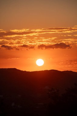 Güneş ufkun altına batıyor, gökyüzünü turuncu ve altın renkleriyle boyuyor. Tepelerin silueti güzel manzaraya derinlik katar..