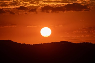 Canlı bir günbatımı, yuvarlanan tepelerin siluetinin üzerine sıcak bir parıltı saçar. Bulutlu gökyüzünün altında huzurlu ve pitoresk bir manzara yaratır..