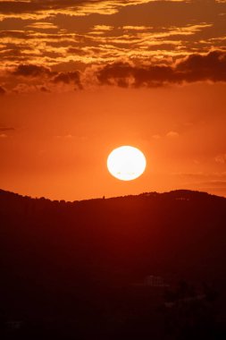 Parlak bir günbatımı, koyu bir sırt çizgisinin arkasında batan büyük bir güneşe ev sahipliği yapar. Gökyüzü, üzerinde bazı bulutlar bulunan ateşli turuncu renklerle doludur..