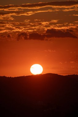 Turuncu gökyüzü ve alçak bulutlarla kızgın bir günbatımı. Güneş, parlak bir ışıltıyla uzak dağların siluetinin altında batıyor..