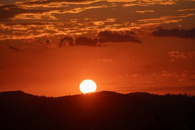 Karanlık tepelerin arkasında parlak bir küre ile gün batımı. Bulutlar gökyüzüne dağılmış, renkli bir manzara yaratıyor..
