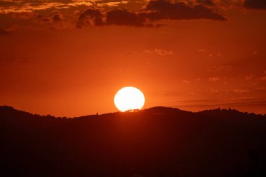 Parlak gün batımı. Güneş kısmen karanlık bir dağ siluetinin arkasında saklı. Turuncu ve kırmızı tonlarda boyanmış bir gökyüzünün altında..