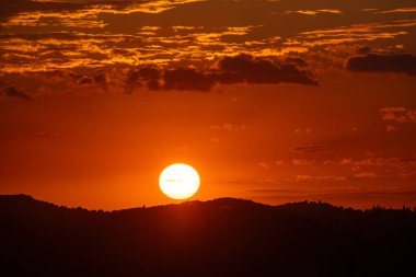 Parlak bir güneş, turuncu gökyüzünün kabarık bulutlarla parıldadığı karanlık ve siluetli bir ufukta batar. Sahne sıcaklığı çağrıştırıyor..