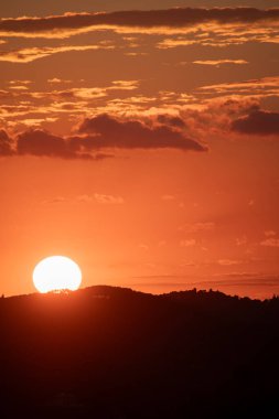 Güneş karanlık, siluetli bir dağ sırasının ardında batarken parlak bir günbatımı, turuncu tonlarda gökyüzünü boyar. Bulutlar doku ekler.