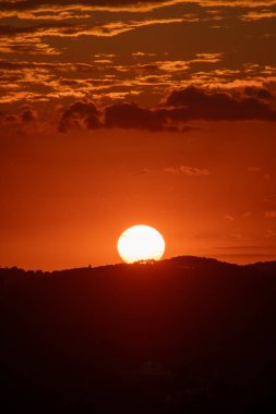 Parlak bir günbatımı gökyüzünü turuncu renklerle boyuyor. Güneş uzak tepelerin siluetinin arkasına batıyor. Güzel bir ekran..
