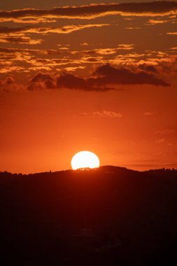 Karanlık bir dağ siluetinin ardında parlak bir güneş batar ve dokulu bulutlarla güzel bir turuncu gökyüzü yaratır. Alacakaranlıkta güzel manzara.