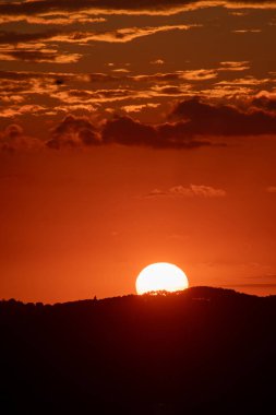 Güneş siluetli bir manzaranın üzerinde batar. Gökyüzü bulutlarla çizilmiş parlak bir portakaldır. Güneş tepenin ardında görünüyor..