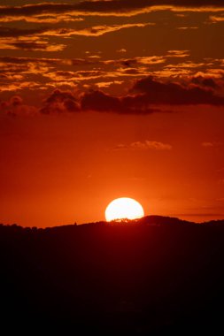 Büyüleyici bir günbatımı sahneye hakim oluyor, sıcak turuncu ve kırmızı bir parıltı saçıyor. Siluetli tepeler dramatik bir ön plan oluşturur.