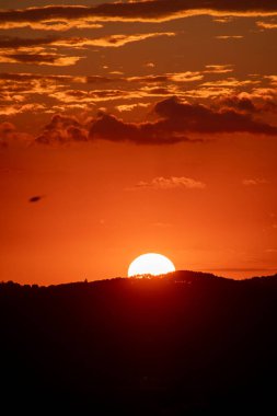 Parlak güneşin karanlık, siluetli bir yamacın üzerinde battığı, bulutların üzerine sıcak bir parıltı saçtığı çarpıcı bir günbatımı sahnesi..