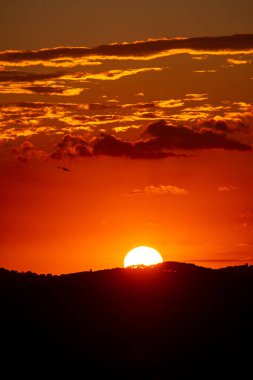 Güneş, gökyüzünde parlak turuncu ve kırmızı renkler ve dağınık bulutlar oluşturan karanlık bir dağ sırasının arkasında batar..