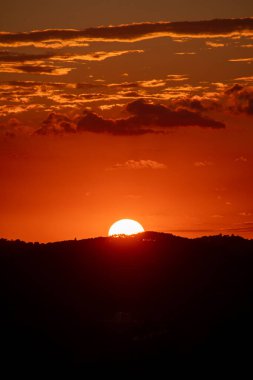 Turuncu ve kırmızı renkleri olan parlak bir günbatımı karanlık ve siluetli bir dağ sırasına inerken ufuk çizgisine hakim oluyor. Bulutlar gökyüzünü dolduruyor.