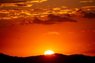Canlı bir günbatımı gökyüzünü turuncuya boyuyor, toprak siluetiyle. Uzakta bir kuş uçar, dingin manzaraya eklenir.