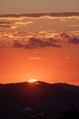 Görüntü canlı bir gün batımı yakalıyor, gökyüzünü dolduran turuncu ve kırmızı renkler, ve siluetli ufuk ve ışığa karşı bina.