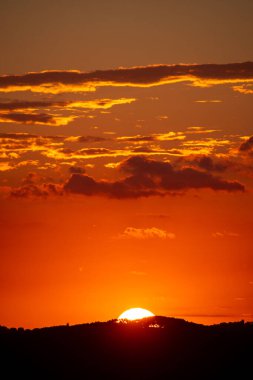 Parlak bir günbatımı gökyüzünü turuncu renkler ile boyar. Güneş ufukta batarken siluet şeklinde yuvarlanan tepeler bulutlar tarafından çerçevelenir..