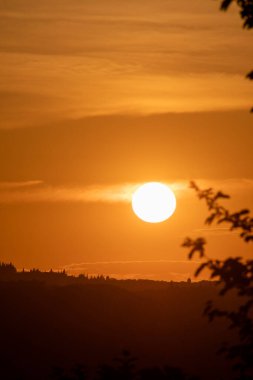 Büyüleyici bir günbatımı, sıcacık bir turuncu ışıltıyla manzarayı yıkıyor. Siluetli ağaçlar ve uzak tepeler derinlik yaratır..  
