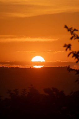 Ufuk çizgisinde kısmen gizlenmiş parlak bir güneş sıcak turuncu renklerde gökyüzünü aydınlatıyor seyrek bulutları ve uzak ağaçları aydınlatıyor..