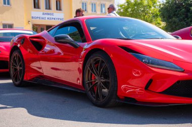 Kırmızı bir performans arabası asfaltta durur. Arka planda bir binanın yanında iki adam duruyor. Sol tarafta kısmen görünür bir araç daha var..