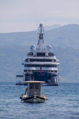 Birden fazla güvertesi, iletişim ekipmanı ve helikopter pisti olan büyük bir yat önünde daha küçük bir servis teknesiyle birlikte suda duruyor..