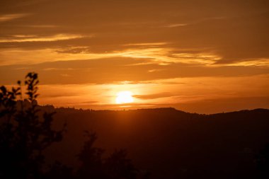 Güneş uzak dağların ardından iniyor, gökyüzüne altın bir parıltı saçıyor. Siluet ağaçları dramatik bir önplan ögesi ekler.
