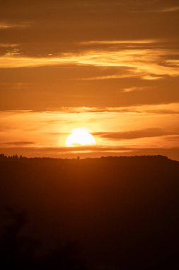 Altın güneş ışığı, güneş uzak bir dağ sırasının üzerinde batarken manzarayı aydınlatır. Gökyüzü ılık renkler ve yumuşak bulutlarla alevler içinde..
