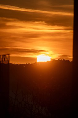 Pencereden yakalanan güneş, turuncu bir alevle, siluet ağaçlarla ve yapraklarla batıyor. Sıcaklık ve sükuneti çağrıştıran sakin bir sahne..