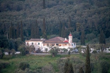 Beyaz binalı bir ev manzarası, tepesinde haç olan bir kule, ve kırsal kesimdeki yüksek ağaçların arasında yemyeşil bir alan..