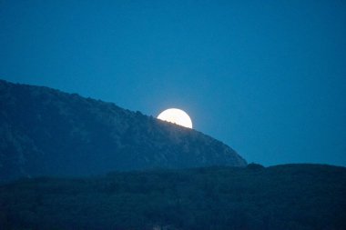 Gece gökyüzünde, dağlık bir arazinin siluetinin arkasında dolunay beliriyor. Sahne maviliklerin gölgesinde ve ay ışıl ışıl parlıyor..