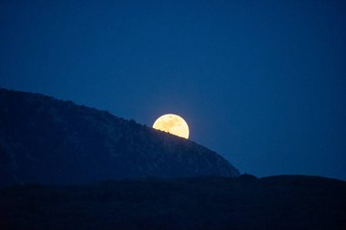 Dolunay, karanlık bir dağ siluetinin üzerinden yükseliyor. Gece atmosferinde çok koyu mavi bir gökyüzünün altında..