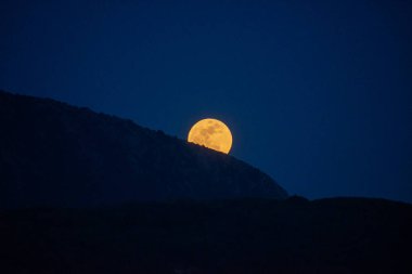 Karanlık bir dağın arkasında parlak bir dolunay parlıyor, mürekkepli gökyüzüne büyüleyici bir parıltı saçıyor. Sahne doğanın ihtişamını çağrıştırıyor..