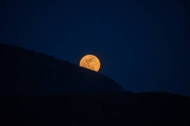 Dolunay, alacakaranlıkta karanlık ve dağlık bir arazinin arkasından yükselir. Gökyüzü koyu mavi, karşıtlık yaratır..