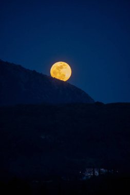 Canlı bir dolunay siluetli bir dağın arkasından yükseliyor, derin mavi gökyüzüne hükmediyor. Ön planda bir kasaba yanıyor..
