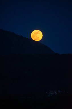 Parlak dolunay yükseliyor, alacakaranlık gökyüzüne karşı karanlık bir dağın arkasında turuncu parlıyor. Gece manzarası huzurlu ve huzurlu..