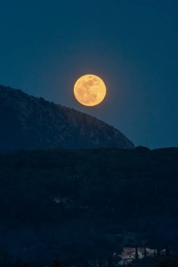 Çarpıcı bir dolunay, karanlık, ormanlık bir dağ ve ay ışığıyla yıkanmış uzak evleri kaplar..