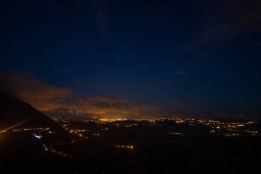 Yükselmiş bir gece manzarası, yıldızlarla ve şehrin ince parıltısıyla benekli bir gökyüzünün altındaki engin bir arazide dağınık ışıkları ortaya çıkarır..