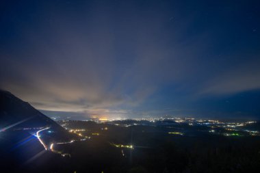 Ele geçirilen, bir dağın arka planına ve bulutlu yıldızlı gökyüzüne gece aydınlatılmış kentsel bir gökyüzünün hava perspektifi..