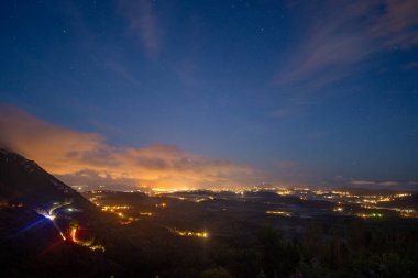Gökyüzünden manzaralı bir gece manzarası, şehir ışıkları parıldıyor, yıldızlı bir gökyüzünde turuncu bulutlar var. Yüksek bir noktadan sakin bir sahne..