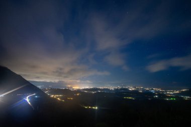 Gece bir kasaba manzarası, bulutlu koyu mavi bir gökyüzünün altında ışıklar parlıyor. Bir dağ kenarda duruyor, yol ışıkları onu takip ediyor.