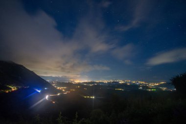 Dağ manzaralı bir gece manzarası. Şehir ışıkları kısmen bulutlu, yıldızlı bir gökyüzünün altında parlak bir şekilde parlıyor. Manzara karanlık ve görkemli..