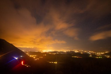 Canlı bir sahne, dramatik bulutlar, yumuşak aydınlatma ve yıldızlı bir gökyüzü ile gece aydınlatmalı bir şehri gözler önüne seriyor..