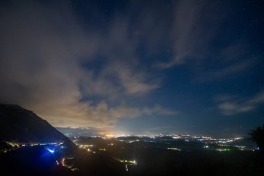 Yüksek bir perspektif, solunda bir dağ ve yıldızlarla dolu bir gökyüzü ve yukarıda bulutlarla dolu bir şehrin gece görüntüsünü yakalıyor..