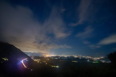 Bir gece sahnesinde yüksek bir dağ manzarasından şehir ışıkları sergileniyor, aydınlık yollar dağdan aşağı sarkıyor yıldızlı bir gökyüzünün altında..