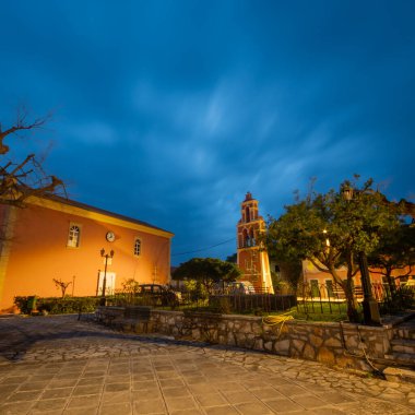 Göz hizasındaki bir fotoğrafta geceleri turuncu bir bina ve gökyüzünün altında çarpıcı bir şekilde aydınlatılmış çan kulesi sergilenen sakin bir plaza yer alıyor..