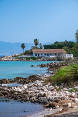 Turkuaz bir sahil boyunca kayalık bir çıkıntı üzerinde, palmiye ağaçları ve yeşilliklerle çerçevelenmiş, kırmızı kiremitli beyaz bir villa bulunur..