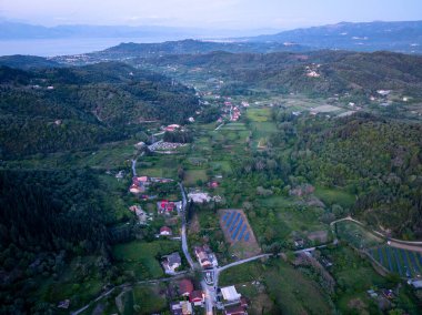Yukardan çekilen yeşil kırsal alanlar evler, ormanlar, tepeler ve bulutlu bir gökyüzü ile buluşan kıyı manzaralarıyla aydınlanır..
