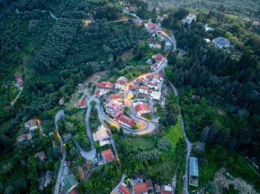Havadan çekilen görüntülerde, yemyeşil ağaçların arasına yerleşmiş, çeşitli mimari yapılara sahip virajlı yollar ve binalar içeren tuhaf bir tepe köyü görülüyor..